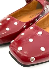 Zapato tipo Mary Jane color cereza charolado, con tachas plateadas, correa con hebilla y suela plana.