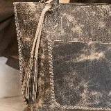 Cartera tipo tote de cuero marrón con costuras artesanales visibles, bolsillo frontal con el logo grabado, asas de hombro y detalles de flecos colgantes.