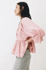 Blusa rosada de algodón con ribetes de encaje, cuello redondo con abertura en V y tiras para anudar, mangas largas abullonadas con puños elásticos y terminación peplum con vuelo.