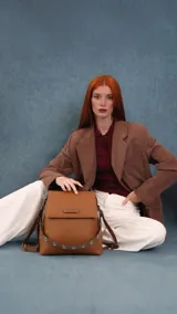 Mochila tipo cartera con tapa, confeccionada en símil cuero color suela. Cuenta con doble compartimento, división interna, cierre de seguridad y bolsillo trasero con cierre. Incluye una correa frontal decorativa con tachas metálicas.