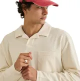 Remera de manga larga tipo polo, color beige, confeccionada en tejido de textura tipo toalla (terry). Presenta cuello camisero, cierre frontal con botones ocultos y un bolsillo aplicado en el pecho.