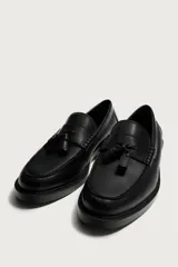 Mocasín de vestir color marrón oscuro, confeccionado en cuero, con detalle de antifaz y borlas decorativas en el empeine. Presenta una horma redonda y suela plana a tono.