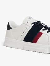 Zapatillas urbanas Tommy Hilfiger de corte bajo, confeccionadas en piel lisa color blanco. Presentan paneles laterales en azul marino y rojo, cordones oscuros y suela de goma gruesa con diseño dentado. Incluyen detalles de la marca en la lengüeta, lateral y talón.
