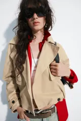 Trench corto de cuello solapa y escote en pico, color beige, con mangas largas terminadas en trabillas con botones. Cuenta con bolsillos laterales con solapa, cierre frontal cruzado con botón y bajo abullonado con elástico interior.