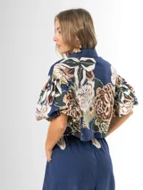 Conjunto de dos piezas confeccionado en viscosa con estampado floral en tonos marrones y verdes sobre base blanca. Incluye una blusa tipo camisa corta con cuello solapa, cierre frontal de botones y mangas abullonadas con diseño globo, y un pantalón tipo culotte de tiro alto, amplio y fluido con pinzas en la cintura.