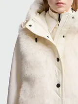 Chaleco Moncler modelo Arve en color blanco crema, con frente y capucha de pelo sintético. Presenta un diseño acolchado con cierre frontal de botones a presión negros, capucha ajustable con cordones y el logotipo de la marca bordado en el pecho.