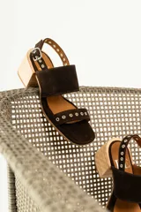 Sandalias de gamuza color marrón con taco cuadrado de madera. Presentan dos tiras anchas, una superior lisa y una inferior decorada con ojales metálicos plateados. Cuentan con cierre de pulsera con hebilla ajustable en el tobillo.