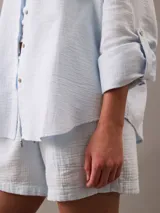 Camisa celeste de gasa doble, con cuello clásico, cierre frontal con botones, bolsillo de parche en el pecho y mangas largas con solapa enrollable.