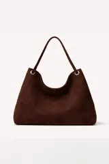 Cartera tipo hobo de gamuza color marrón oscuro, con asa única y detalles de argollas metálicas plateadas en los extremos.