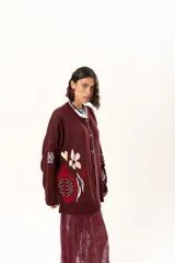 Saco de punto color vino con diseño holgado, cuello redondo y mangas largas. Presenta un gran bordado central en la espalda con forma de flor en tonos crema y rojo, acompañado de un pequeño bordado de sol y una frase bordada en la parte inferior.
