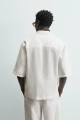Camisa de lino color blanco, con cuello de solapas de muesca, manga corta y cierre frontal cruzado oculto. Presenta un calce relajado y aberturas laterales en el bajo.