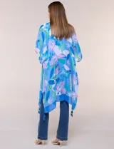 Kimono de gasa azul con estampado botánico de hojas tropicales en tonos celestes, verdes y rosados. Presenta un diseño abierto al frente con mangas cortas y amplias, ruedo asimétrico estilo pañuelo y detalle de borlas en las esquinas inferiores.