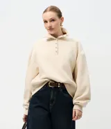 Buzo de algodón color beige, con cuello polo y cierre frontal parcial con botones dorados. Presenta un diseño de corte holgado con mangas largas y puños acanalados.