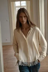 Camisa de satén color crema con cuello solapa, manga larga y cierre frontal con botones ocultos. Presenta un detalle de encaje en el bajo asimétrico.
