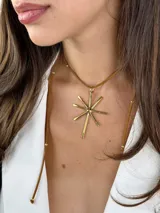 Colgante de diseño minimalista compuesto por una estructura circular rígida de metal dorado y un dije central en forma de estrella o destello de líneas rectas. Incluye tiras de cuero marrón con detalles metálicos que aportan un toque artesanal.