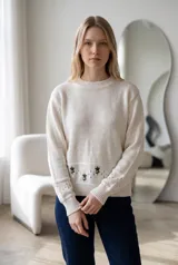 Sweater de punto color crema con cuello redondo, manga larga y diseño de calados en los puños. Presenta un detalle de bordado floral en la parte inferior y una franja con perforaciones decorativas.