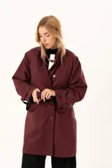 Trench de corte holgado color bordó, con cuello camisero, cierre frontal con botones y cinturón ajustable con hebilla. Presenta mangas largas con detalles de correas en los puños y bolsillos laterales.