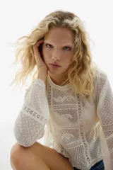 Blusa de manga larga confeccionada en malla plumeti color crema, con detalles de encaje, bordados y alforzas. Presenta cuello redondo, cierre con botón en la nuca y un corte con frunces laterales en la cintura que aportan volumen al bajo.