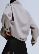 Chaqueta gris de diseño corto y holgado, con cuello alto envolvente, mangas anchas y puños ajustables con tiras y botones. Cuenta con bolsillos laterales y cierre frontal oculto.