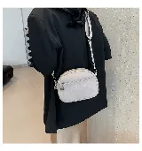 Cartera bandolera color gris claro, de cuero sintético, con correa ancha de tela estampada y cierre superior con cremallera.