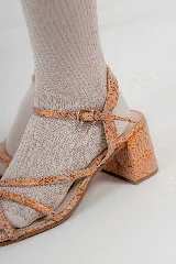 Sandalia de cuero metalizado color piel con detalles en naranja, con plantilla cuadrada y taco de 5 cm. Presenta tiras finas cruzadas en el empeine y una tira ajustable con hebilla en el tobillo.