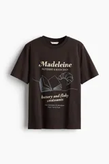 Remera marrón de corte recto con cuello redondo y mangas cortas. Estampado frontal con el texto "Madeleine PASTISSERIE & BOOK SHOP", el dibujo de un libro abierto, un croissant y la frase "buttery and flaky croissants".