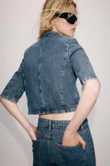 Blazer cropped de denim azul con cuello subido, manga corta y bolsillo de vivo en el pecho. Cierre frontal con botón.