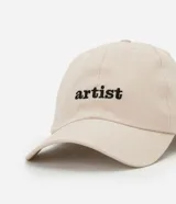 Gorra de sarga color beige con visera curva y bordado frontal con la palabra "artist" en negro. Ajuste trasero con hebilla metálica.