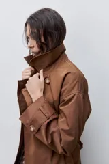 Trench clásico color marrón, de silueta relajada, confeccionado en tejido liviano, con cuello de solapa, doble abotonadura frontal y mangas largas con presillas regulables en los puños.