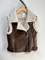 Chaleco estilo gamulán de cuero sintético marrón con interior y solapas de corderito sintético color crema. Presenta cierre frontal cruzado, bolsillos laterales con cierre y correas ajustables en los laterales del ruedo.