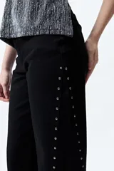 Pantalón de denim corte wide leg color marrón, con ruedo desflecado y detalle de tachas metálicas a lo largo de las costuras laterales.