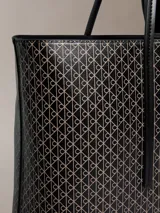 Cartera tote Calvin Klein de diseño estructurado, color negro y marrón con estampado de monograma CK en toda la superficie. Cuenta con asas de mano dobles y finas, y viene acompañada de una mini cartera lisa con cierre y detalle del logo de la marca en dorado.