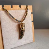 Collar con cadena de cuentas facetadas en tonos dorados y grises, con dije en forma de colmillo con detalles esmaltados en verde y figura dorada.
