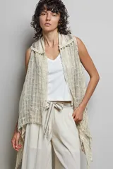 Cardigan sin mangas confeccionado en algodón con tejido texturizado y bajo asimétrico. Presenta bordes deshilachados, bordados detallados y adornos de monedas doradas a lo largo de las solapas.