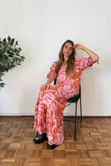 Vestido largo con estampado de hojas tropicales en tonos naranja y rosado. Presenta escote en V, mangas cortas abullonadas con ajuste de lazo y falda con volados en el ruedo.