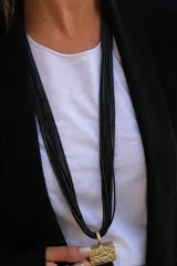 Collar largo de múltiples hilos negros con dije rectangular de textura rugosa en tono dorado y fleco largo de hilos negros colgante.