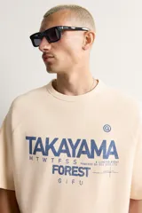 Camiseta relaxed fit de color beige, con cuello redondo, mangas cortas y estampado frontal en tono azul con texto gráfico.