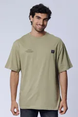 Remera oversize color verde oliva con cuello redondo, mangas cortas y un parche cuadrado negro con un logo en el pecho. Presenta un estampado de texto en el lado izquierdo del pecho.