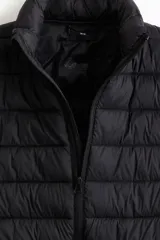 Chaleco acolchado negro de nylon con diseño guateado, cuello elevado, cierre frontal con protección para el mentón y bolsillos laterales. Corte entallado y diseño repelente al agua.