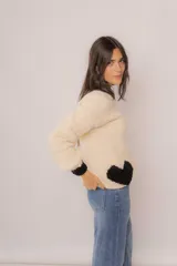 Buzo tejido a mano en lana merino, color crema con detalles en negro en el cuello, puños y un diseño de corazón en la parte inferior. Presenta un corte amplio y mangas largas.