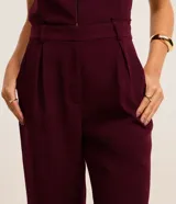 Pantalón zanahoria color borgoña de crepé de poliéster, con trabillas en la cintura, cierre tradicional, pliegues y bolsillos.