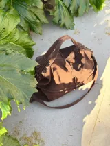 Bolso de cuero marrón con diseño tipo morral, cuenta con doble correa de hombro y correa larga ajustable, cierre superior y detalles de remaches metálicos.