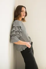 Blusa de tela con estampado vichy en blanco y negro, con cuello redondo y mangas con tres volados escalonados que llegan hasta el codo. Presenta puños elásticos y cierre con botón en la parte posterior.