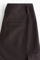 Pantalón cargo de sarga color marrón oscuro, con tiro alto y corte holgado de piernas tipo barrel con pinzas, bolsillos de fuelle con solapa a los lados y trabilla abotonada en el bajo.