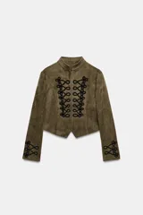 Chaqueta corta de antelina color verde militar, con cuello subido, manga larga y bajo asimétrico. Presenta detalles de alamares negros bordados en contraste en el frente y en los puños.