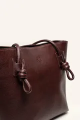 Cartera tote de cuero suave color marrón, con diseño amplio y líneas simples, con tiras de hombro decoradas con nudos artesanales y pequeño logo grabado en el centro.