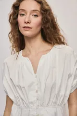 Vestido corto de algodón color blanco, con escote en V y botonadura frontal. Presenta mangas cortas holgadas, cintura ajustada con frunces y falda acampanada con terminación de volado en el ruedo.