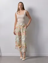 Pantalón capri de lino color crudo con estampado de hojas en tonos verdes, amarillos y naranjas. Presenta bolsillos laterales y detalle de puntilla en el ruedo.