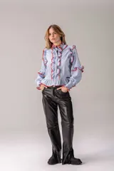 Camisa celeste con volados decorativos en los hombros, pecho, mangas y ruedo, destacados con ribetes en tono rojo. Presenta cierre frontal de botones, cuello alto y mangas largas abullonadas con puños ajustados.
