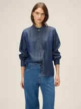 Camisa de denim liviano color azul, con cuello mao y detalles de volados fruncidos en el cuello y la botonera frontal. Presenta mangas largas con puños rectos y corte relajado.
